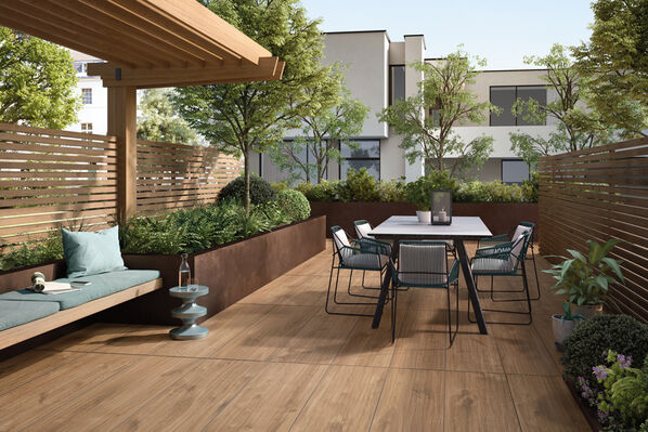 Große, längliche Terrasse, deren Boden mit Holzfliesen (Villeroy & Boch Oak Park OUTDOOR 20) ausgekleidet ist. In der Mitte steht ein Tisch mit Stühlen. Umgeben ist die Terrasse von hölzernen Sichtschutzelementen und grünen Pflanzen. 