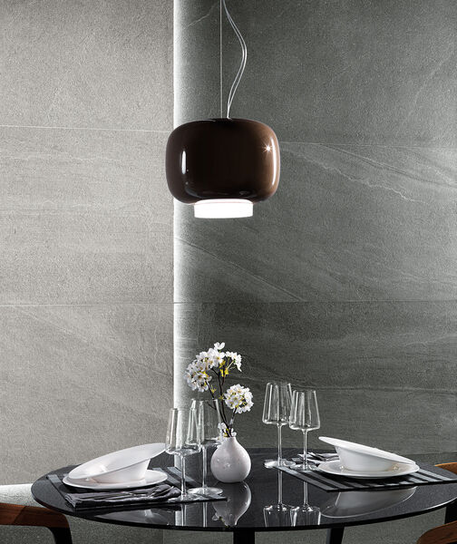 kleiner runder und gedeckter Tisch, über dem eine Lampe hängt. Dahinter eine Wand in grauen Fliesen, in Steinoptik gefliest. 