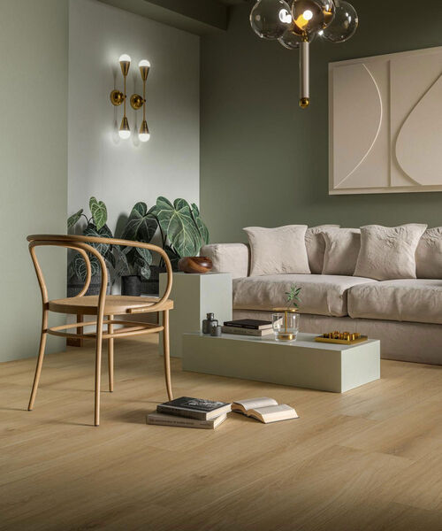Gemütliches Wohnzimmer, in dem eine Couch, ein Stuhl und ein kleiner Tisch zu sehen sind. Der Boden in Holzoptik ist gefliest mit der Marazzi Oltre in der Farbe Sand und gleicht einem eleganten Parkettboden. 