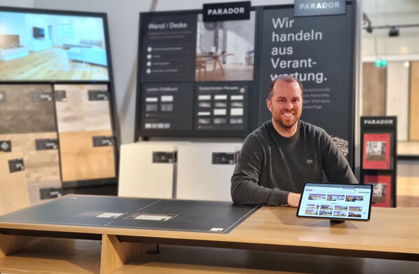 In einem Ausstellungsraum für Echtholzboden sitzt der Geschäftsführer, Tobias Aussieker an einem Schreibtisch und lächelt in die Kamera.