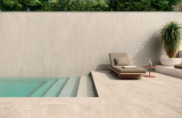 Terrasse mit Pool und Liege. Der Terrassenboden und die abgrenzende Wand sind gefliest mit Fliesen in Natursteinioptik (Novabell Deepstone),