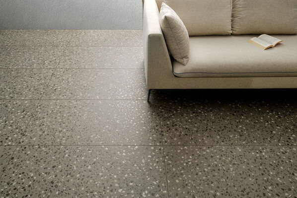 Fliesenboden in dunkler Terrazzooptik, auf dem ein helles Sofa angeschnitten zu sehen ist: Bei der Fliese handelt es sich um die Coem Terrazzo.